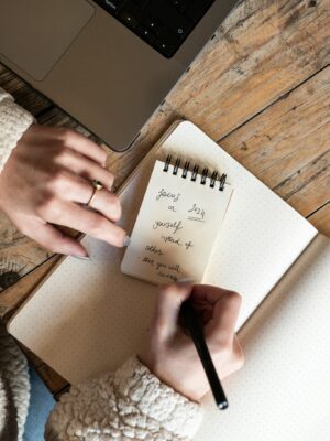 Top view of a woman writing notes in a notepad next to a laptop on a wooden table.