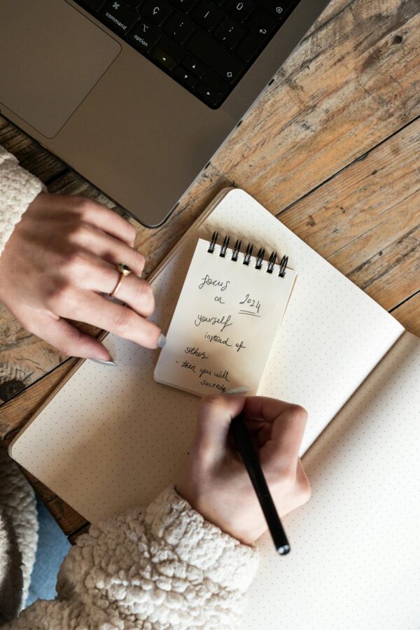 Top view of a woman writing notes in a notepad next to a laptop on a wooden table.