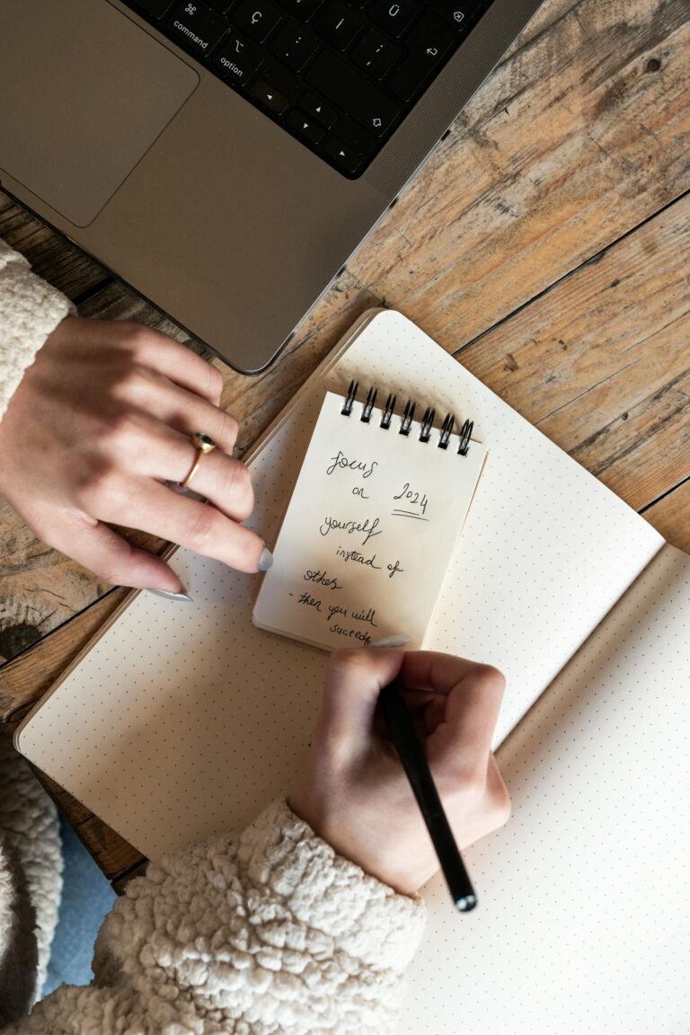 Top view of a woman writing notes in a notepad next to a laptop on a wooden table.