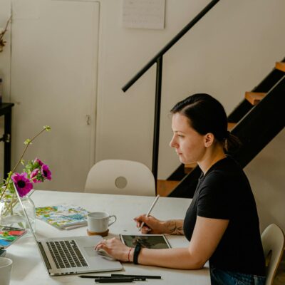 Woman using a digital pen with her laptop in a creative workspace featuring art materials.